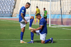Two soccer players in blue jerseys on a green pitch; one kneels to tie the other’s boot near the goal.