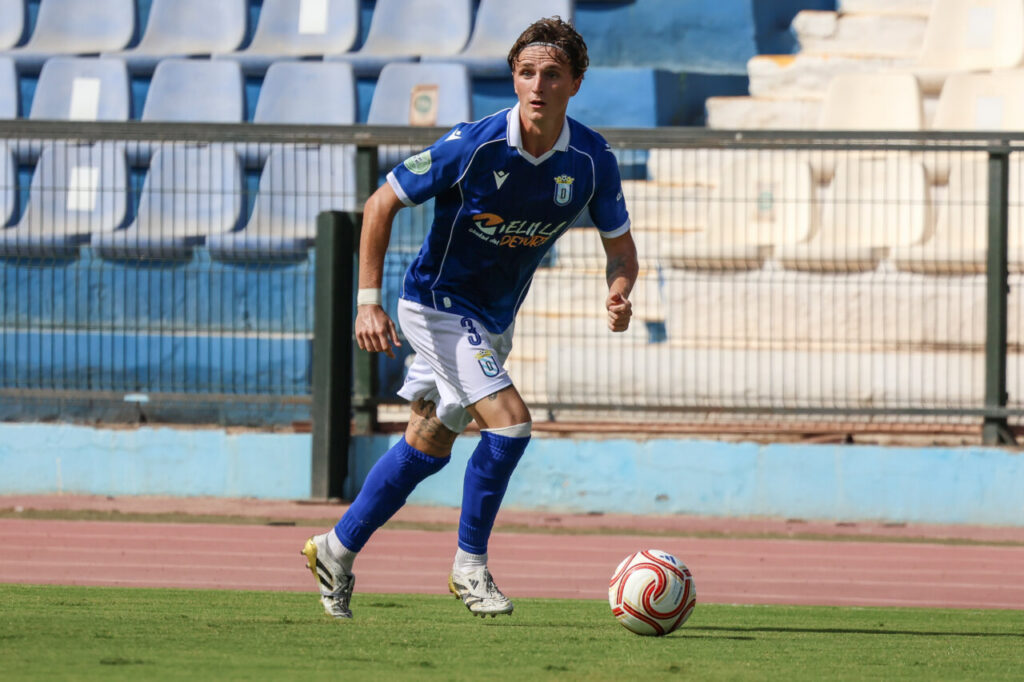 Soccer player in a blue jersey and white shorts dribbles a ball on a grass field with empty stadium seats in the background.