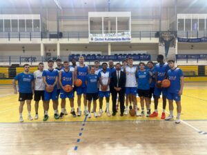 Group of basketball players posing for a team photo on an indoor court; coach in a suit stands center.
