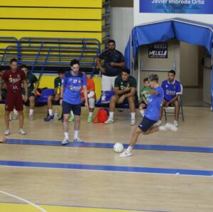 Indoor futsal game: a player in blue kicks a ball while teammates and substitutes watch from the bench and sidelines in a gym with yellow seating.