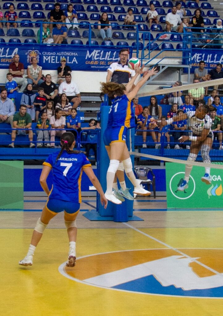 Jugadoras disputan una acción de voleibol junto a la red ante espectadores.