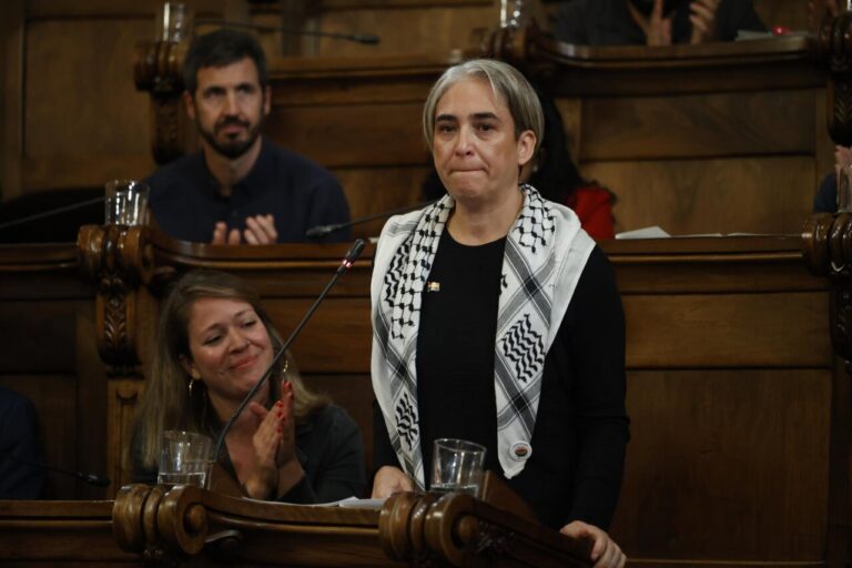 Ada Colau de pie ante un micrófono en una sala con bancos de madera.