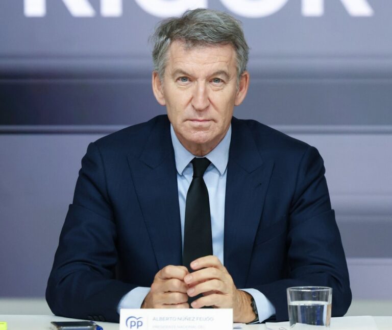 Middle-aged man in a dark suit and black tie sits at a press conference table, hands clasped, with a glass of water nearby.
