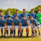 Once futbolistas posan en fila sobre un campo de fútbol con uniforme azul y verde.