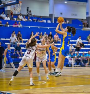 Female basketball player in blue jumps to take a shot near the basket while defenders in white attempt to block, indoors with spectators watching.