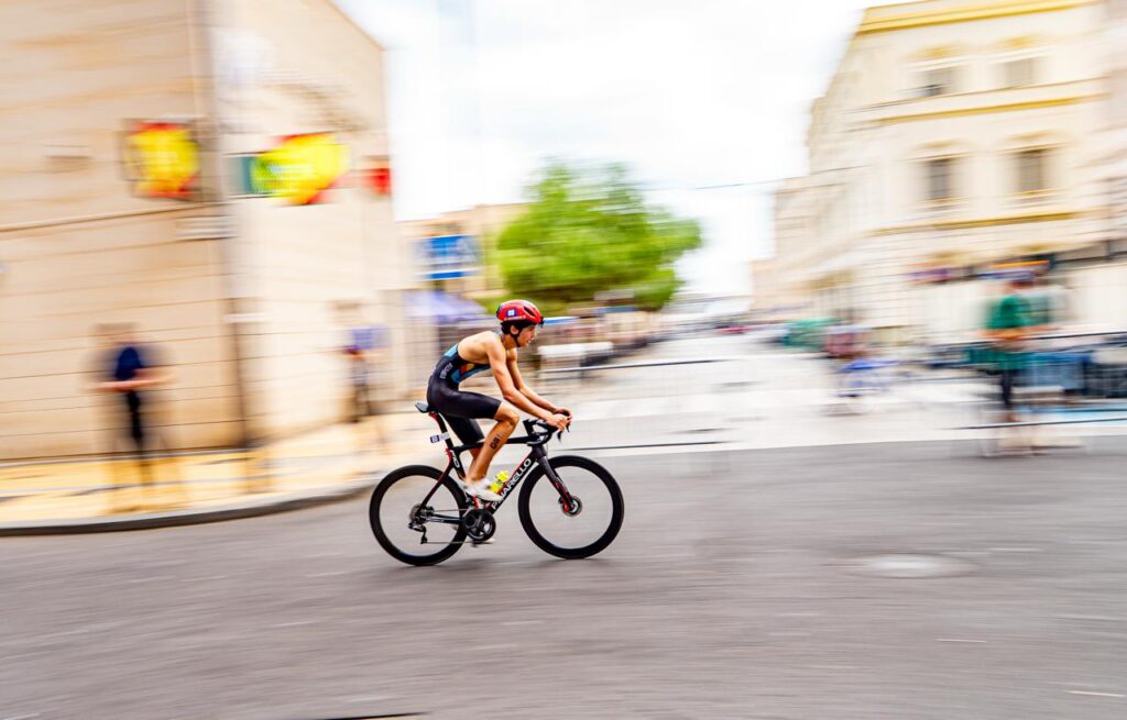 Un ciclista con casco avanza en bicicleta por una calle, con edificios desenfocados.