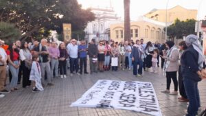 Crowd of men, women, and children standing in a circle around a large banner on the pavement during a peaceful demonstration in a sunny plaza with buildings in the background.