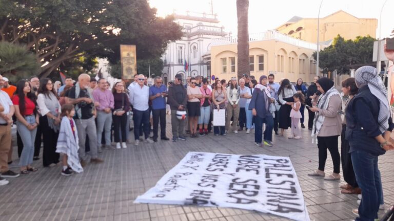 Crowd of men, women, and children standing in a circle around a large banner on the pavement during a peaceful demonstration in a sunny plaza with buildings in the background.