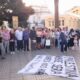 Crowd of men, women, and children standing in a circle around a large banner on the pavement during a peaceful demonstration in a sunny plaza with buildings in the background.