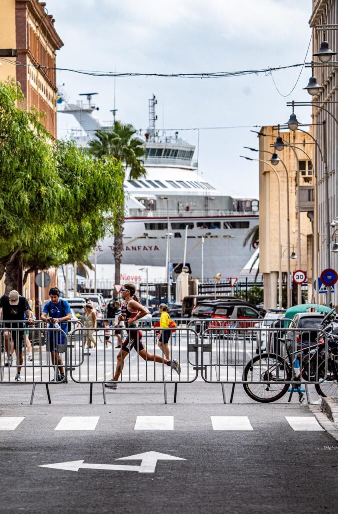 Runners race along a barrier-lined city street with a large white ship docked in the port behind them.