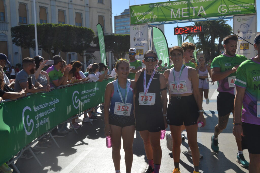 Runners crossing the finish line of a charity race in Melilla, with cheering spectators behind green barriers and a timer reading 00:24:24.