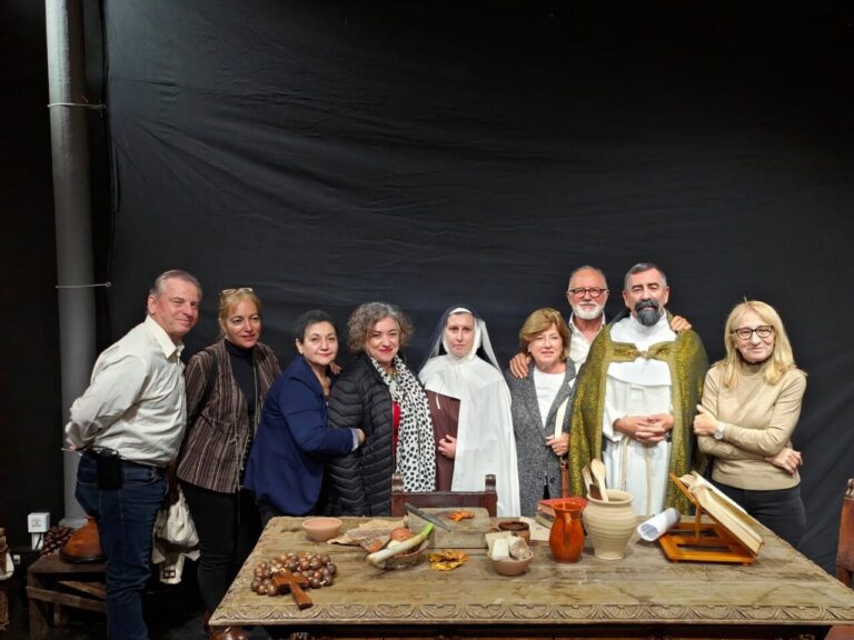 Ten people pose together behind a carved wooden table with pottery, bread, and religious items; a priest in gold vestments stands with the group.