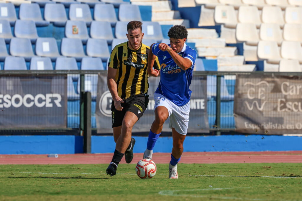 Un futbolista azul disputa el balón con un jugador de negro y amarillo.
