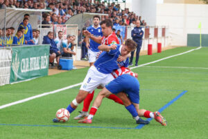 Futbolistas de azul y de camiseta rojiblanca disputan el balón junto a la banda