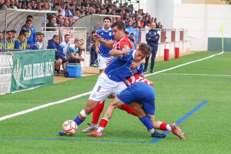 Futbolistas de azul y de camiseta rojiblanca disputan el balón junto a la banda
