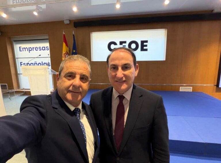 Two businessmen in suits take a selfie in a conference room, with a blue stage and a screen reading 'CEOE' behind them and Spanish and EU flags nearby.