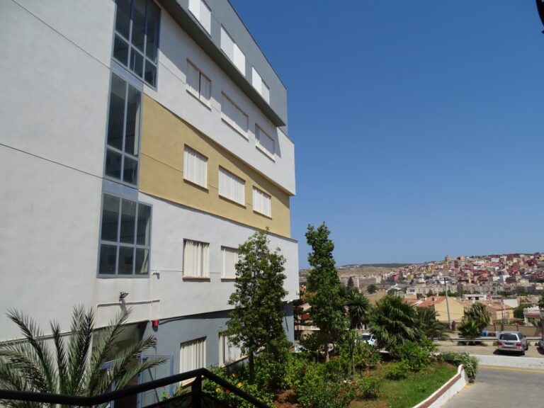 Edificio de fachada blanca y ocre junto a árboles y viviendas bajo cielo despejado