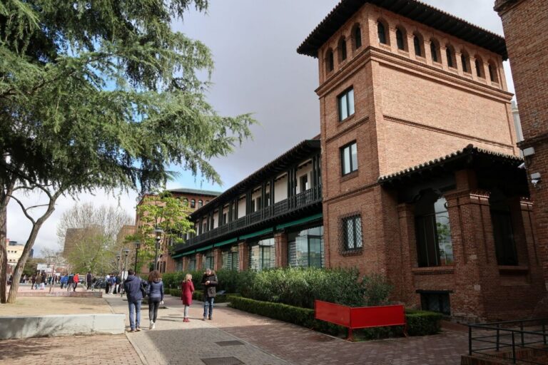 Edificio de ladrillo con galerías junto a un paseo arbolado y varios peatones.