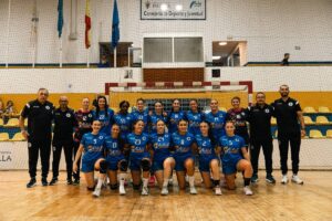 Group photo of a women's volleyball team in blue uniforms posing with coaches inside a gym, in front of a goal net and yellow seats.