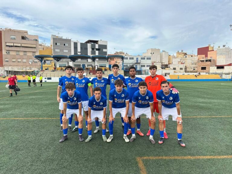 Group of young soccer players in blue uniforms posing on a turf field before a match, with a goalkeeper in orange in the background.L