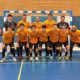 Eleven-player futsal team in orange jerseys posing in front of a goal on an indoor blue court.