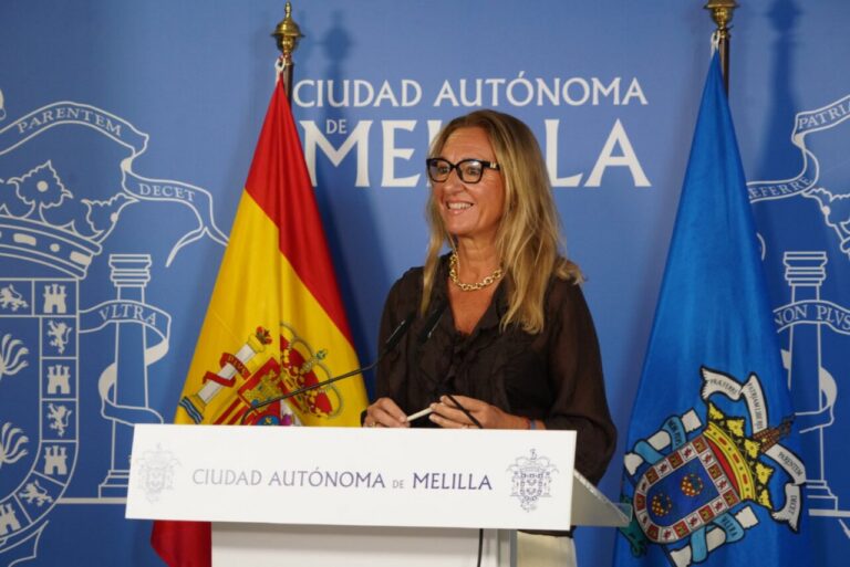 Woman with glasses speaks at a podium labeled Ciudad Autónoma de Melilla, with Spanish and blue Melilla flags behind her.