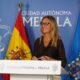 Woman with glasses speaks at a podium labeled Ciudad Autónoma de Melilla, with Spanish and blue Melilla flags behind her.