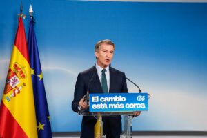 A man in a dark suit speaks at a podium with a blue sign saying 'El cambio está más cerca', flanked by the Spanish and EU flags against a blue backdrop.