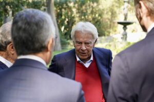 Felipe González conversa con varios hombres en un jardín, vestido con jersey rojo y chaqueta azul.