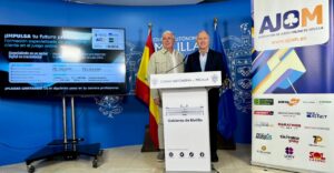 Dos hombres junto a un atril de Melilla, con pantallas y cartel de AJOM
