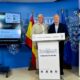 Dos hombres junto a un atril de Melilla, con pantallas y cartel de AJOM