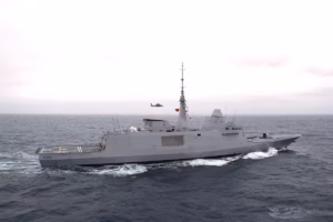 Grey military destroyer cruising at sea with a helicopter flying nearby above the deck area of the ship's stern.
