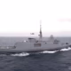 Grey military destroyer cruising at sea with a helicopter flying nearby above the deck area of the ship's stern.