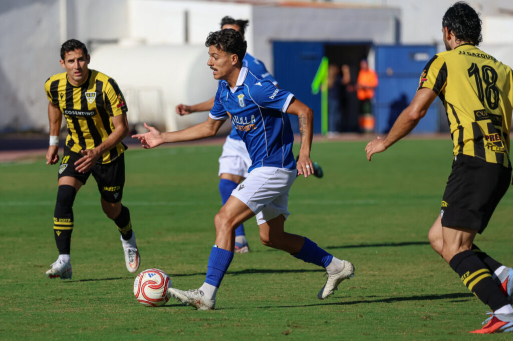 Un jugador de azul conduce el balón ante dos rivales de amarillo y negro.