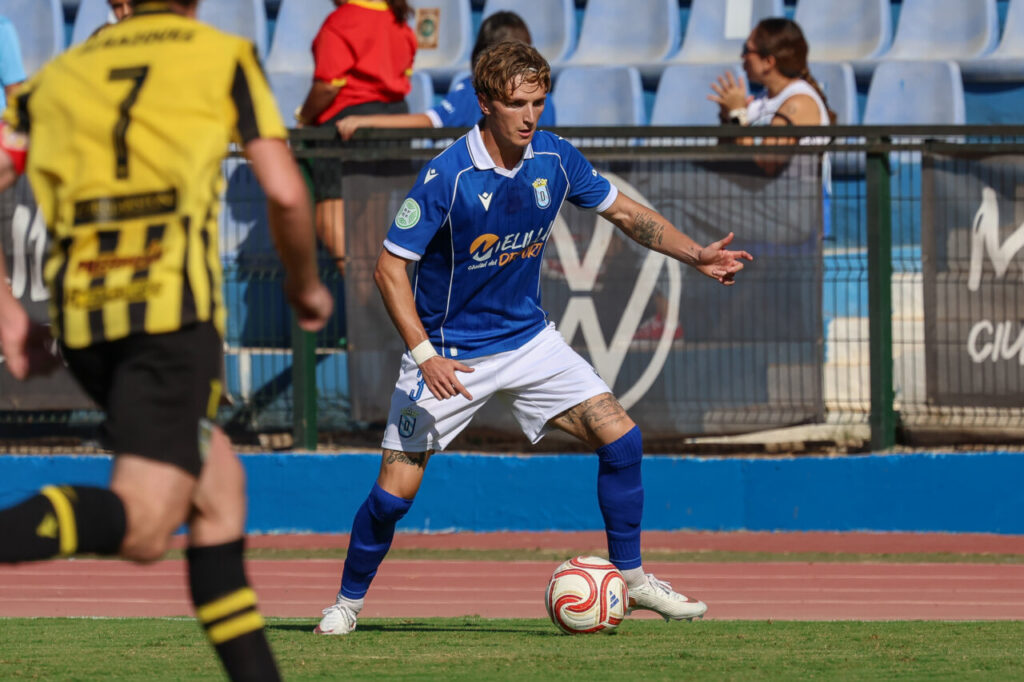 Soccer player in blue kit dribbles the ball on a sunny field, with a yellow-jersey opponent in the foreground.