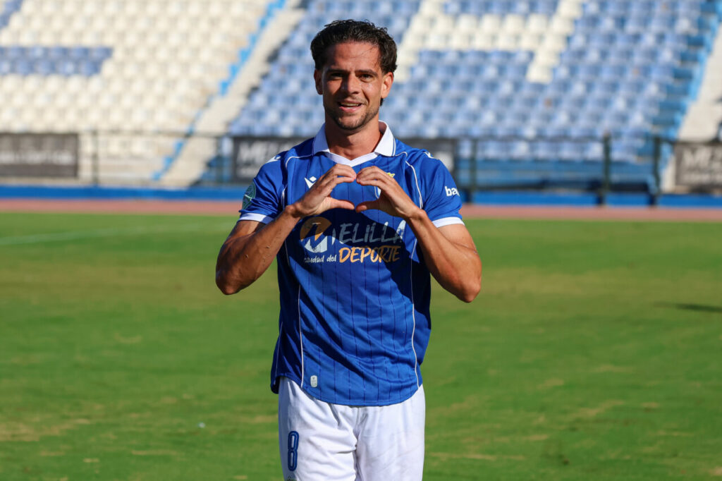 Male soccer player in a blue jersey on a green field, forming a heart shape with his hands at the stadium.