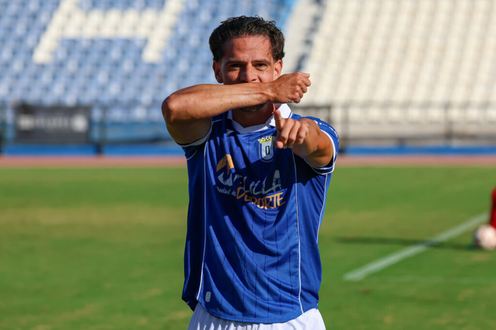 Futbolista con camiseta azul señala hacia delante sobre el césped del estadio.