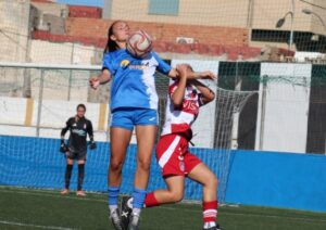 Dos futbolistas disputan un balón aéreo durante un partido en un campo de césped artificial.