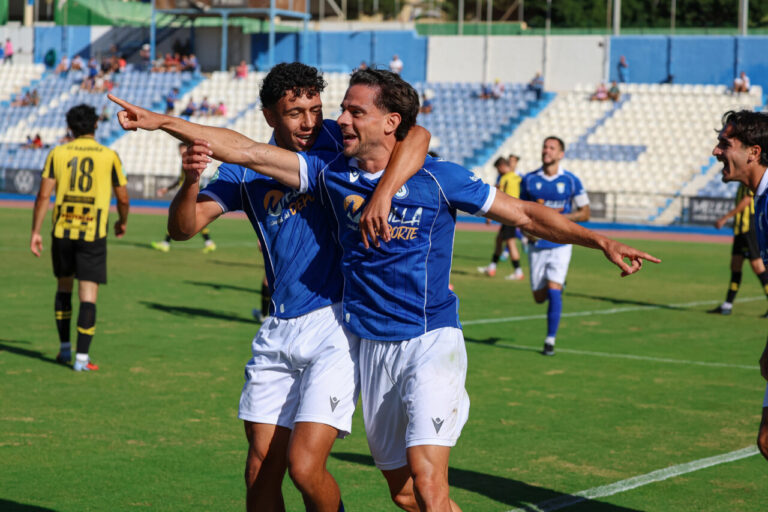 Dos futbolistas con uniforme azul celebran juntos y señalan en el campo