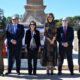 Seis personas posan frente a un monumento, con un militar uniformado a la izquierda.