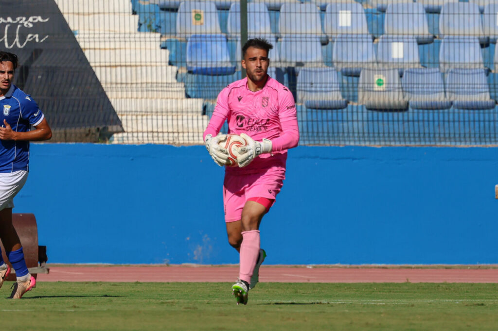 Guardameta con uniforme rosa sostiene un balón mientras un jugador azul aparece parcialmente a un lado.