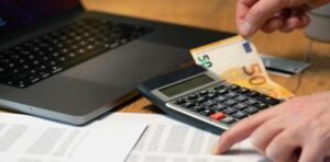 Person using a calculator beside a laptop with euro banknotes on a desk, budgeting or calculating expenses.