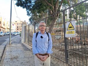 Hombre frente a una verja junto a árboles y un cartel de paso de camiones
