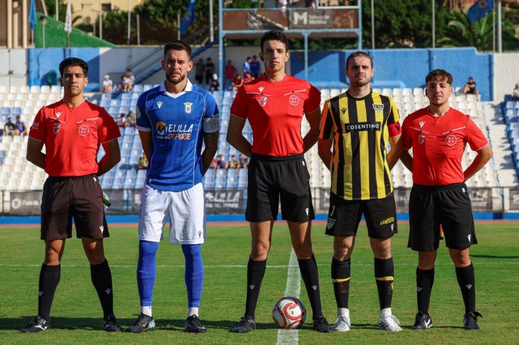 Five soccer players stand in a line on a grass field before a match, wearing red, blue, and yellow-striped uniforms with a soccer ball at their feet.