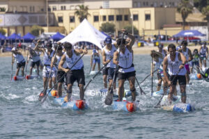 Team of stand-up paddleboarders racing on the water, paddling in unison with tents and a beach backdrop in the background.