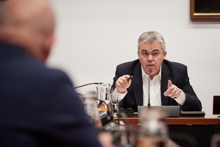 Man in a dark suit speaks into a microphone at a formal meeting, gesturing with a pen in hand.