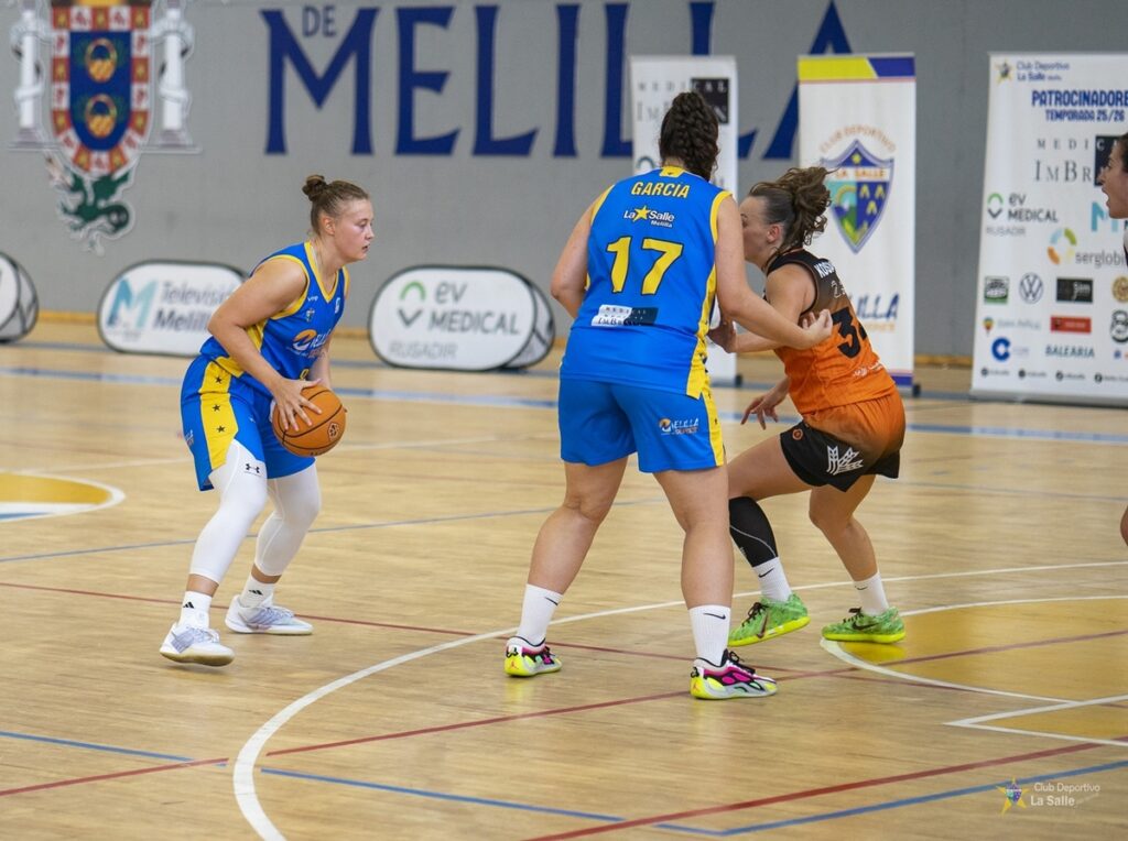 Una jugadora azul y amarilla sostiene el balón mientras una rival naranja la defiende.