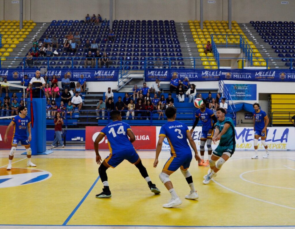 Jugadores de voleibol disputan una jugada en una pista cubierta con gradas al fondo