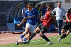 Un jugador de azul disputa el balón con un rival vestido de rojo.