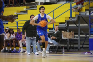 Jugador de baloncesto con uniforme azul avanza con el balón ante el banquillo.